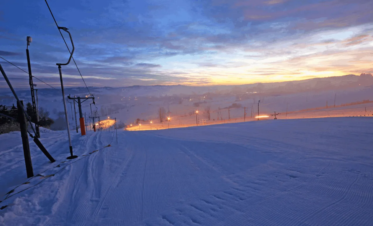 Ośnieżony stok narciarski na Olczańskim Wierchu o wschodzie słońca, z wyciągiem krzesełkowym i latarniami.
