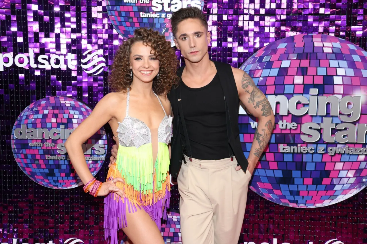 Ola Filipiak, w wieku X lat, w kolorowej sukience z frędzlami, pozuje z partnerem na tle błyszczącej ścianki 