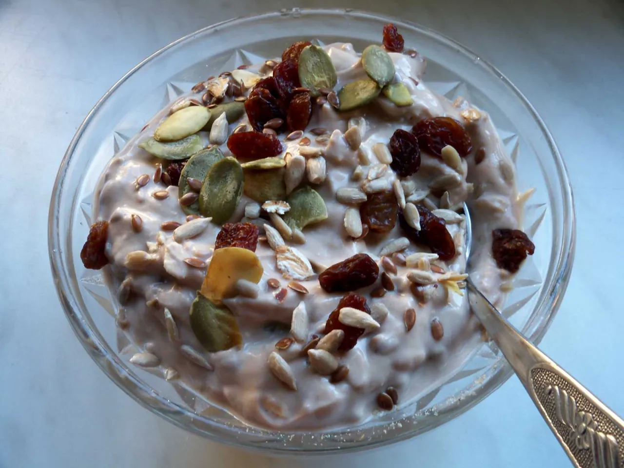 Jak zrobić dietetyczne musli? Miska pełna jogurtu z płatkami owsianymi, pestkami dyni, słonecznika, siemieniem lnianym i rodzynkami.