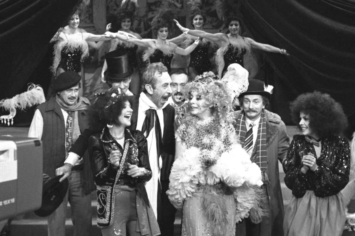 Artyści kabaretu Olgi Lipińskiej w barwnych strojach, wśr&oacute;d nich panie w pi&oacute;rach i panowie w kapeluszach, tworzą radosną atmosferę.