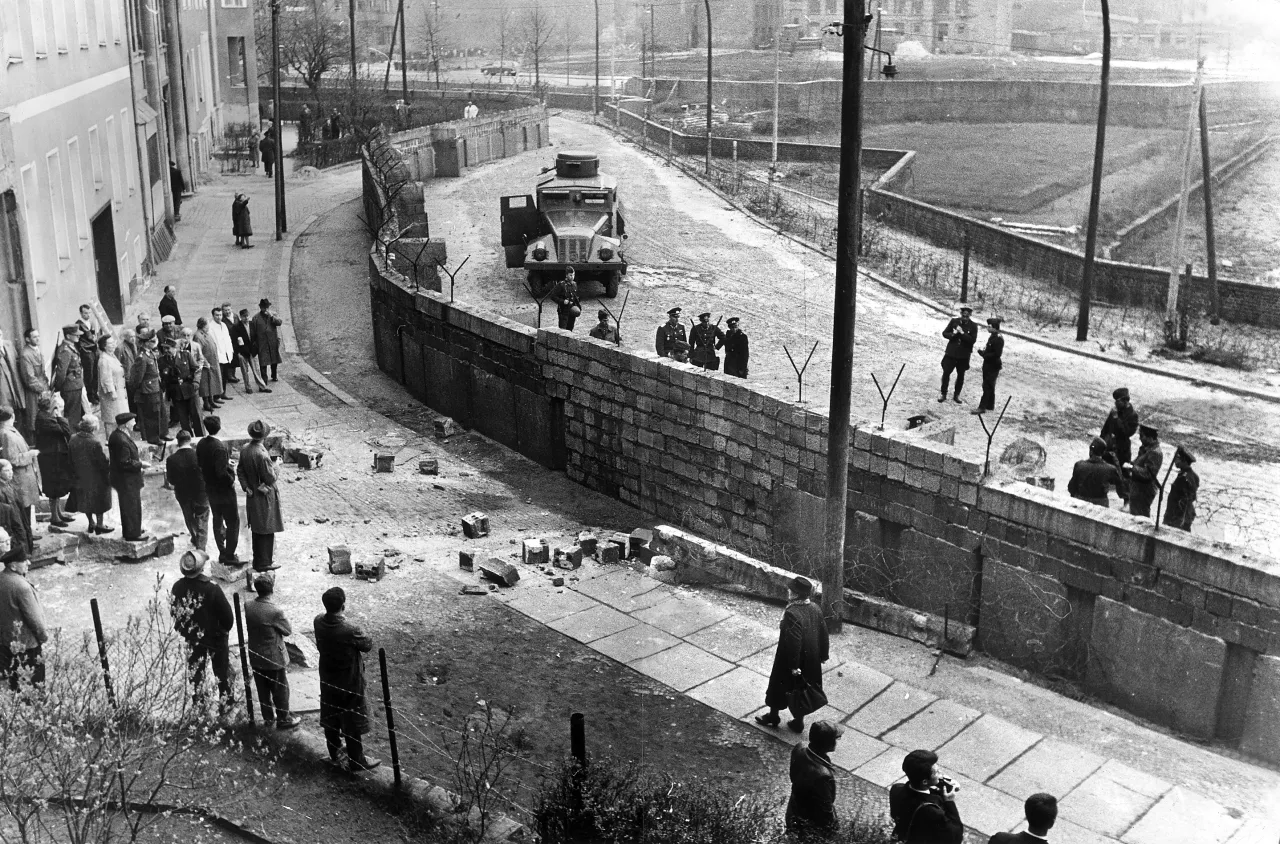 Scena z czasów, kiedy powstał mur berliński. Żołnierze pilnują granicy, a obok stoją zaciekawieni mieszkańcy.