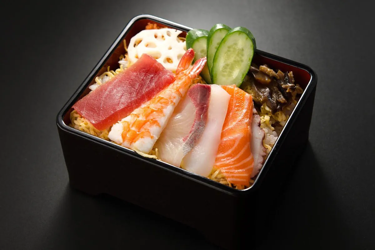 Chirashi zushi: kolorowe plastry ryb (łosoś, tuńczyk, biała ryba), krewetka, plasterki og&oacute;rka i kwiat lotosu na ryżu.