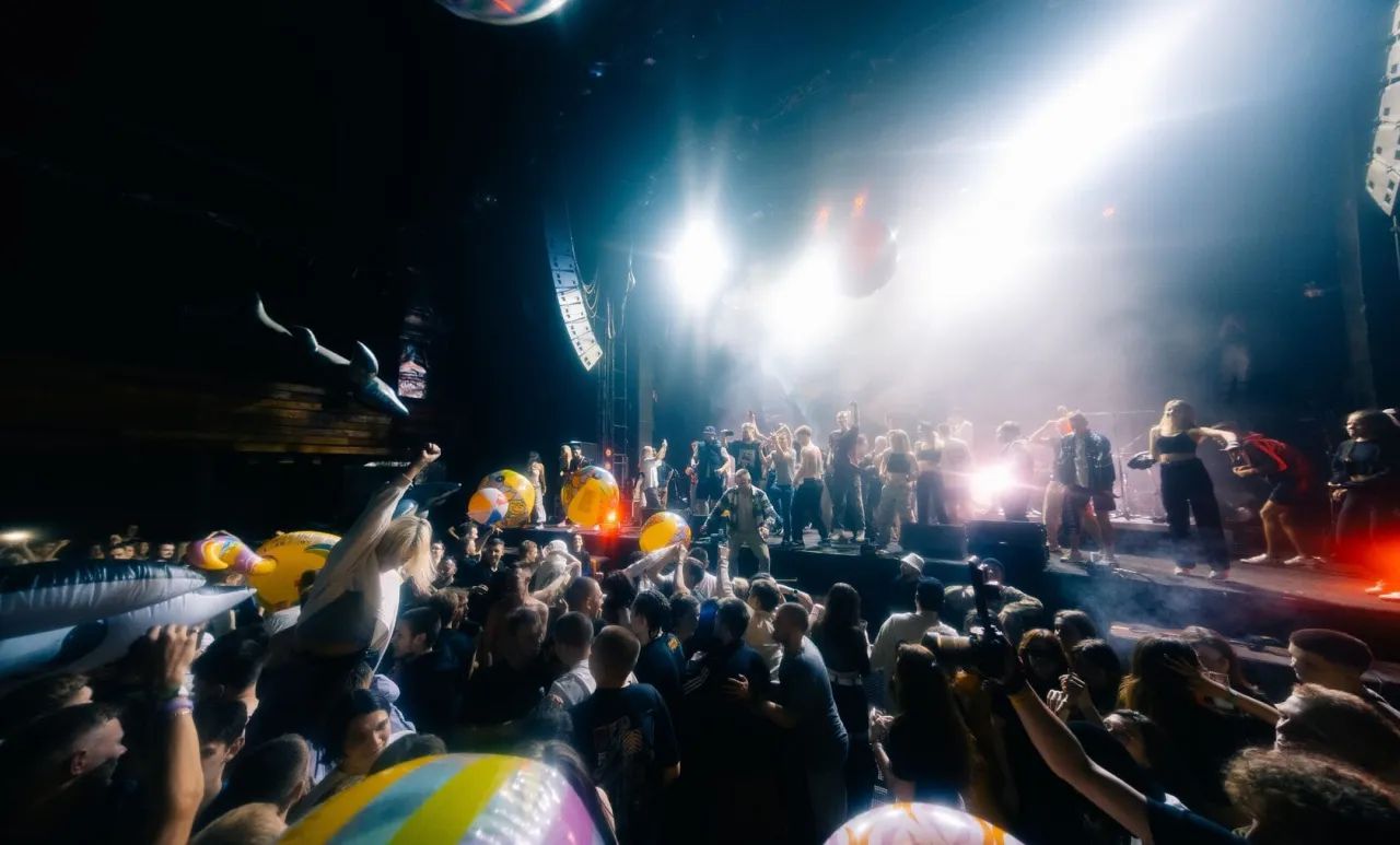 Tłum szaleje na koncercie, rzucając dmuchanymi rekinami i piłkami. Wszyscy zastanawiają się, ile trwa koncert Maty.