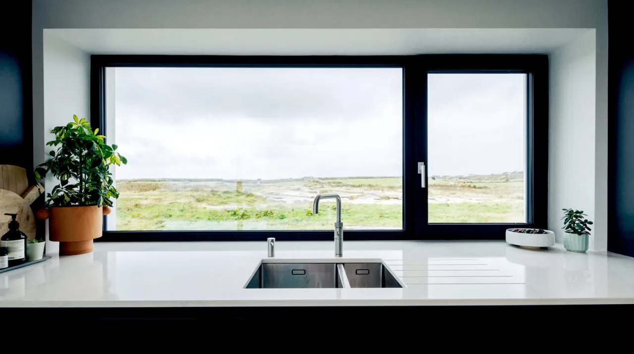 Nowoczesna kuchnia z podwójnym zlewozmywakiem i baterią pod dużym oknem, z którego roztacza się widok na zielone pola. Okna w kuchni są na wysokości blatu.