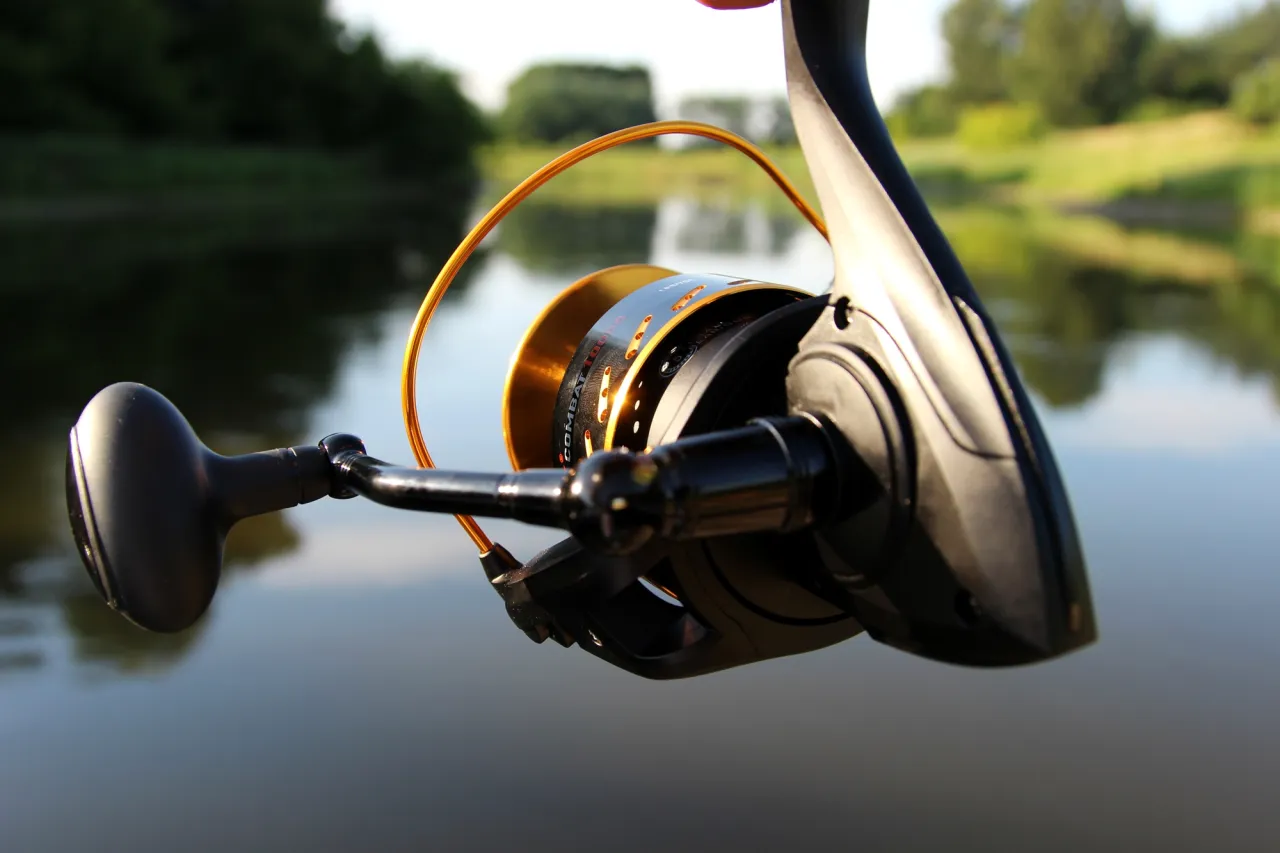 Jaki kołowrotek do spinningu na szczupaka? Ten złoty kołowrotek z czarnymi detalami, idealny na drapieżniki, prezentuje się na tle spokojnej wody i zielonych brzeg&oacute;w.
