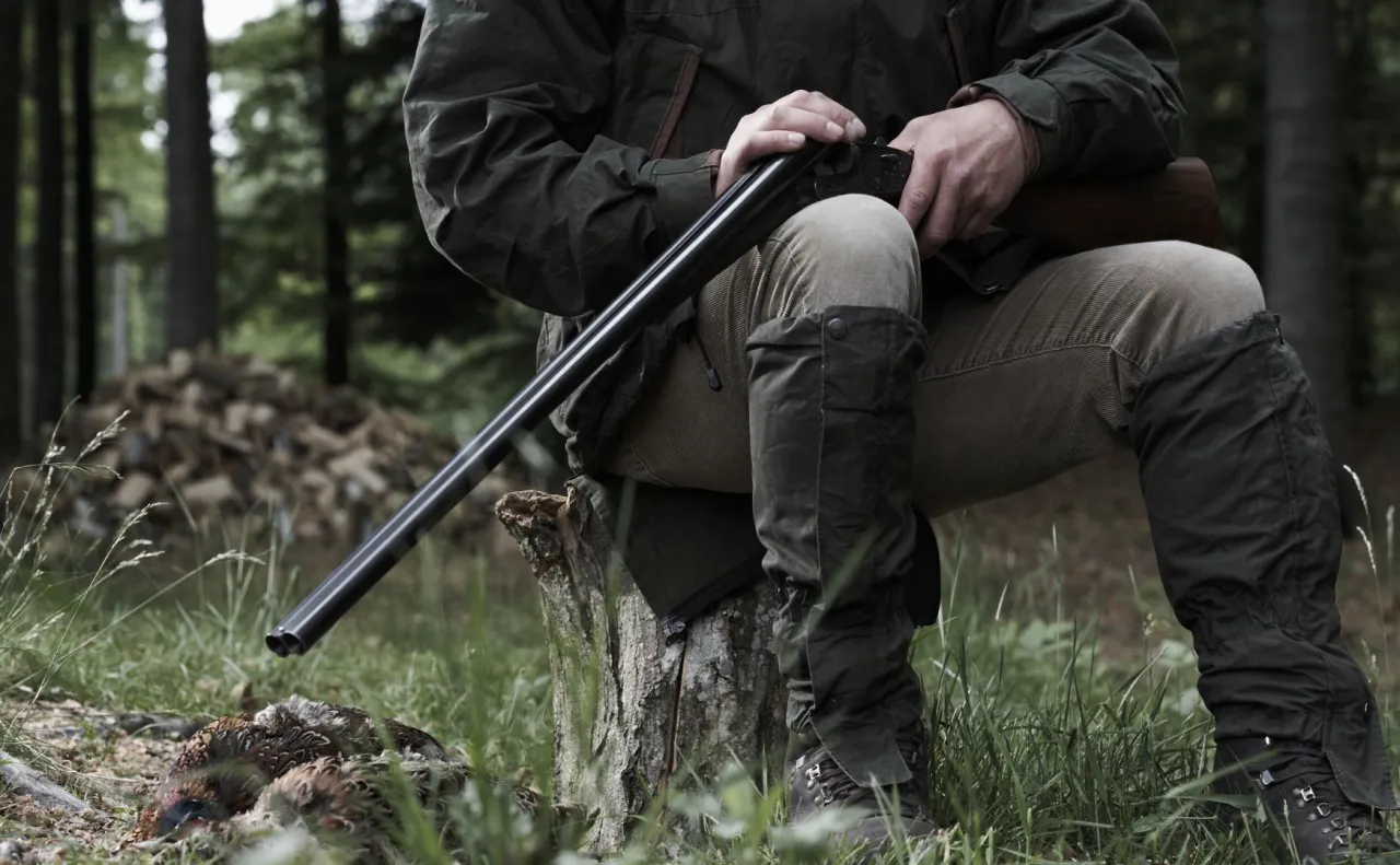 Myśliwy siedzi na pniu, trzymając strzelbę. Obok leży upolowany ptak. Takie chwile to esencja tego, co mogą myśliwi.