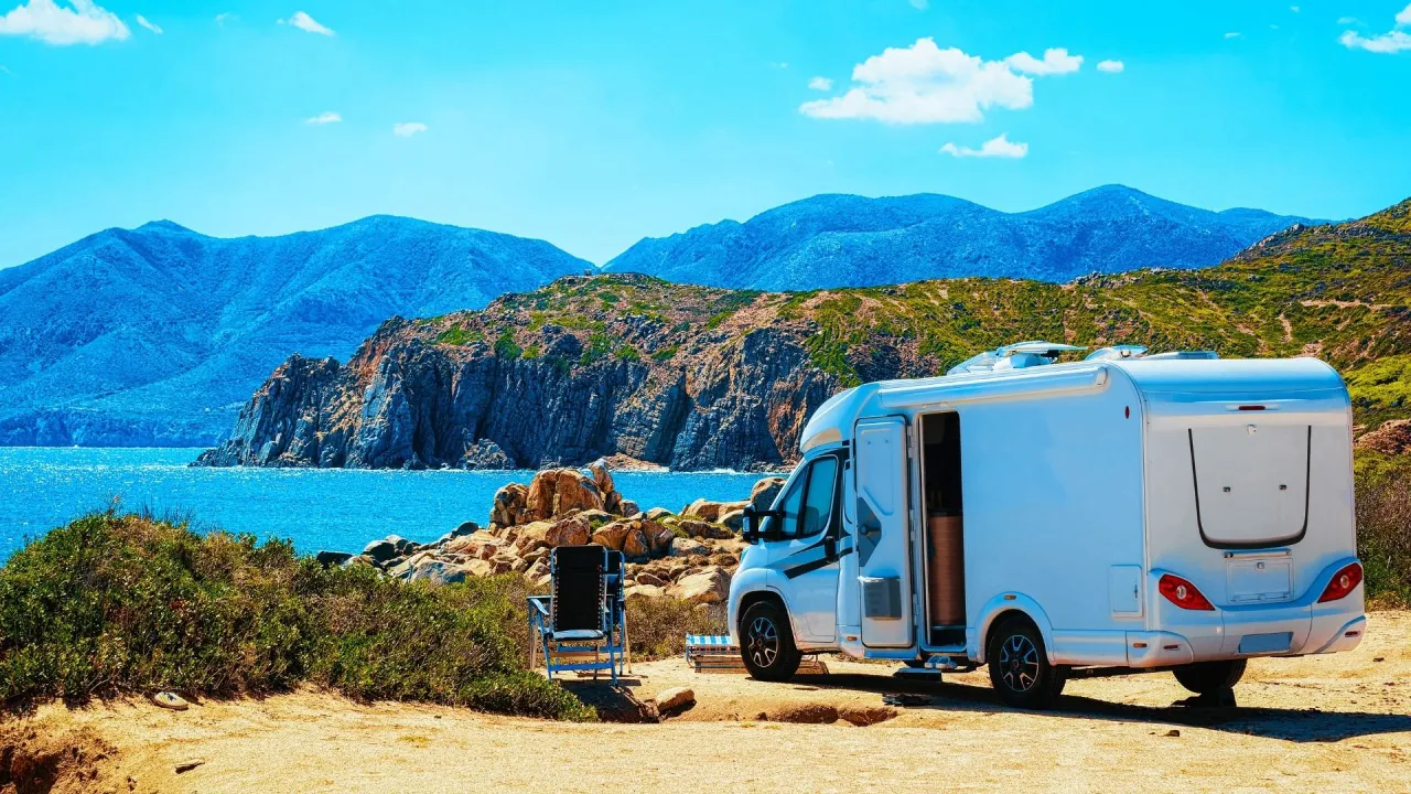 Biały kamper zaparkowany na skalistej plaży, z widokiem na turkusowe morze i g&oacute;ry. Idealne miejsce na podr&oacute;ż kamperem do Włoch.