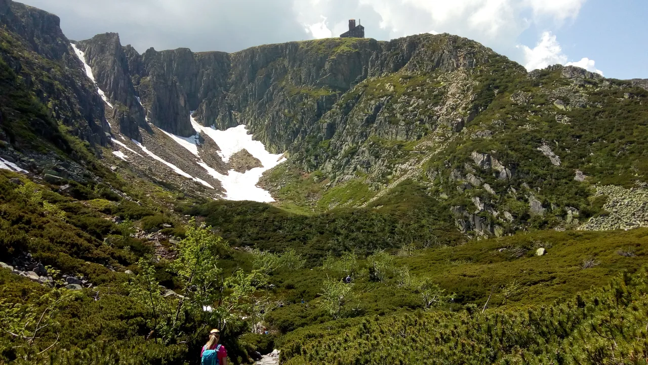 Widok na góry z resztkami śniegu, skalnymi zboczami i zieloną roślinnością. Na szczycie widać schronisko pod Łabskim Szczytem.