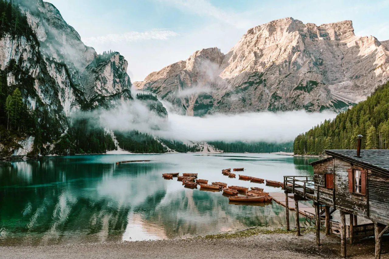 Spokojne jezioro w Tyrolu we Włoszech, otoczone mglistymi g&oacute;rami i lasem. Na wodzie kołysze się ł&oacute;dź, a przy brzegu stoi drewniana chata.