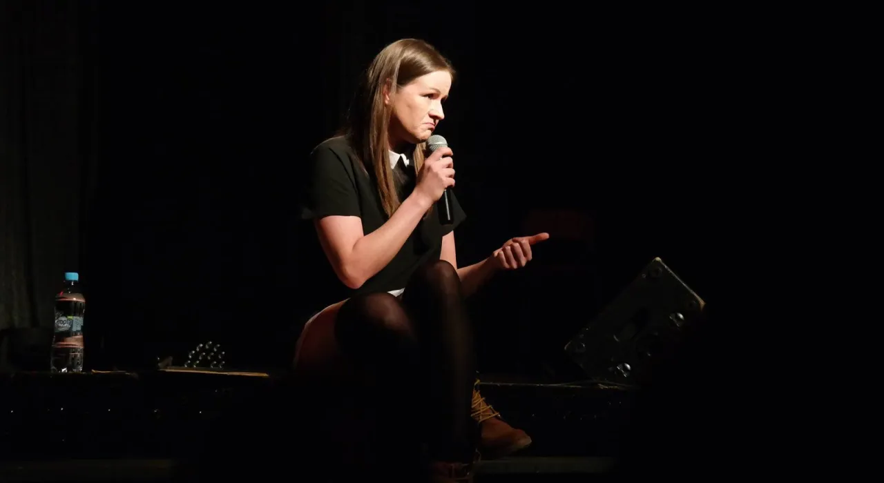 Ewa Błachnio, znana z kabaret&oacute;w, siedzi na scenie z mikrofonem, gestykulując.