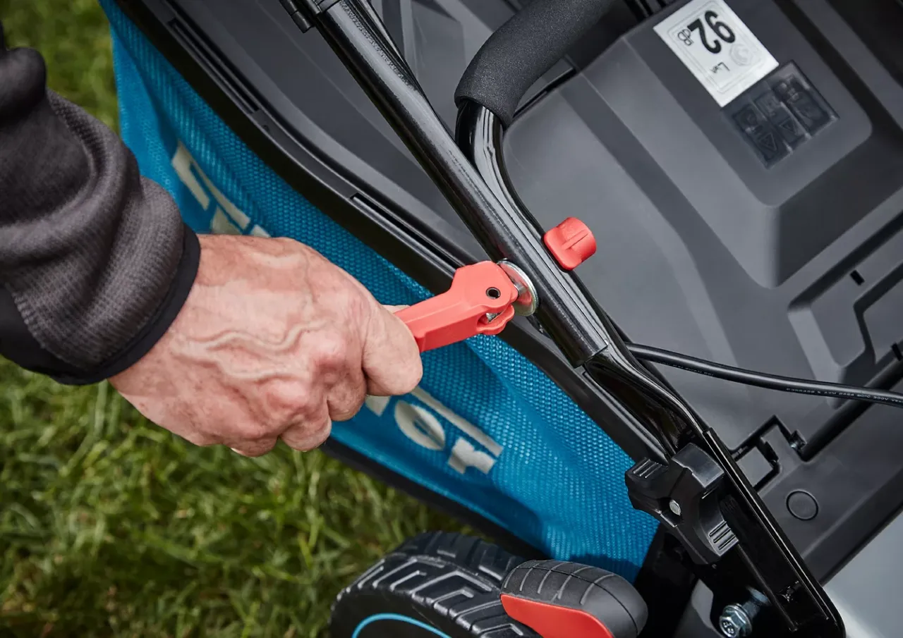 Dłoń chwyta za czerwony uchwyt linki gazu, przygotowując się do odpalenia kosiarki spalinowej.