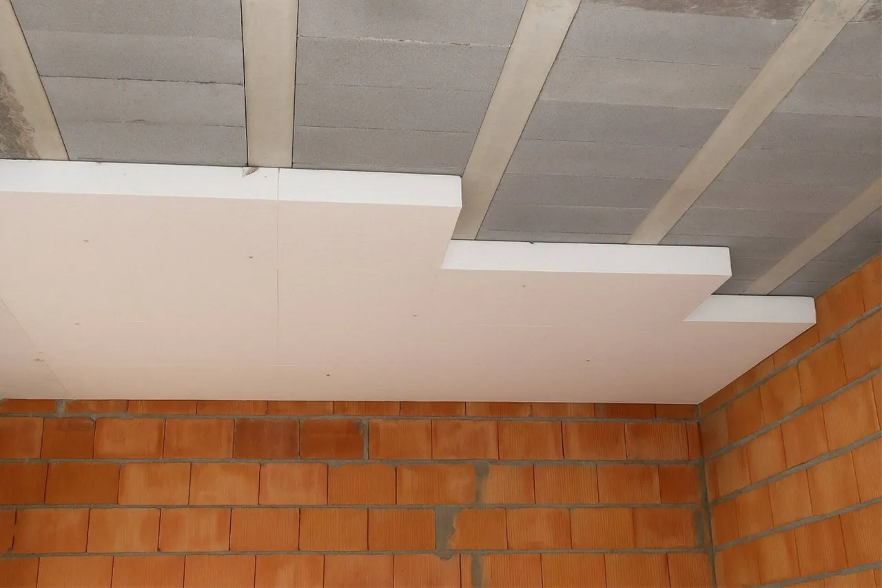 Ocieplanie stropu styropianem. Białe płyty styropianowe montowane pod betonowym stropem, widoczne ceglane ściany.