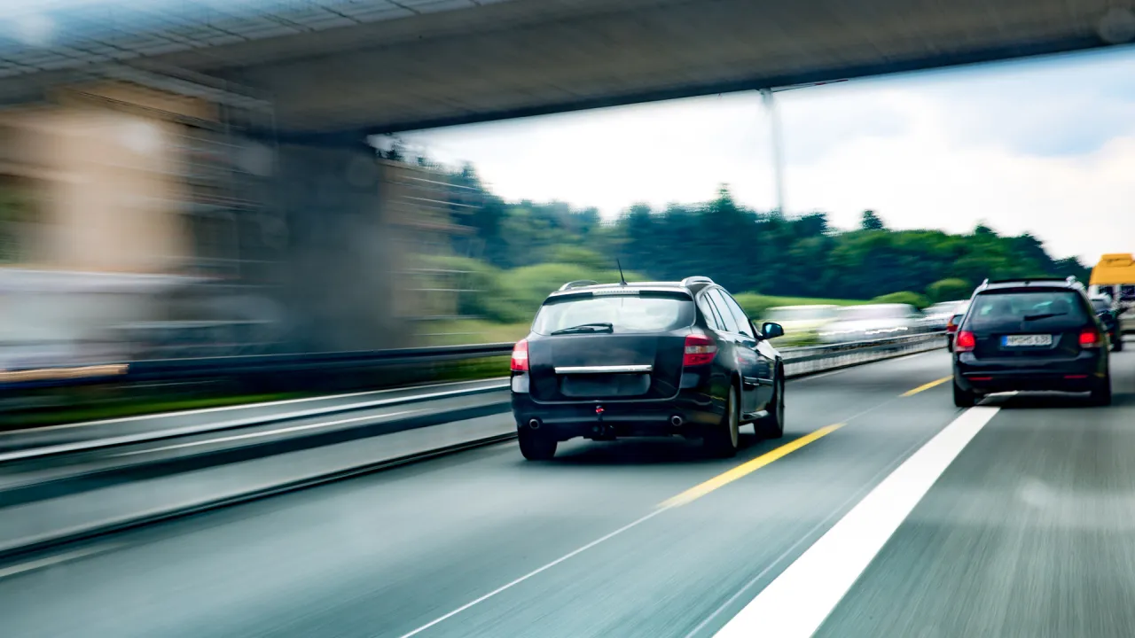 Czarny kombi jedzie autostradą pod wiaduktem. Inne samochody w ruchu. Uważaj, by nie dostać mandatu za zbyt wolną jazdę.