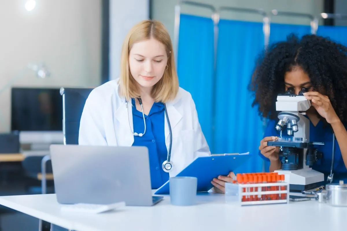 Dwie lekarki pracują w laboratorium. Jedna analizuje próbki pod mikroskopem, druga notuje dane. Czy praca w laboratorium jest trudna? Tak, wymaga skupienia i precyzji.