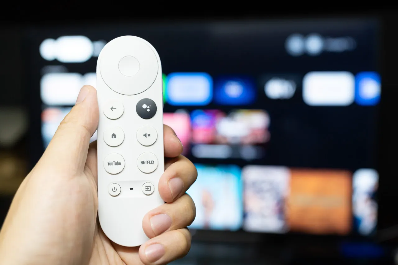 Dłoń trzyma biały pilot z przyciskami YouTube i Netflix. Ekran telewizora w tle pokazuje menu aplikacji, sugerując, jak zaktualizować Chromecast.