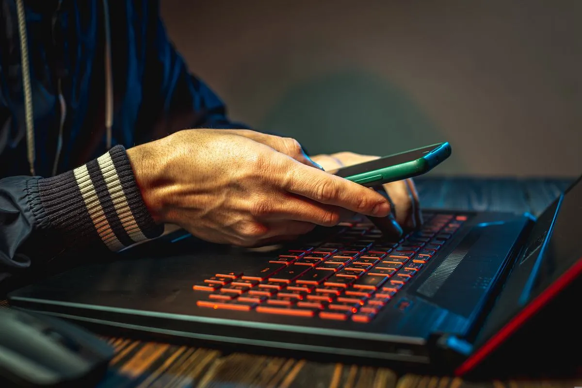 Ręce piszą na podświetlanej klawiaturze laptopa, trzymając telefon. Dowiedz się, gdzie zgłosić oszustwo internetowe.