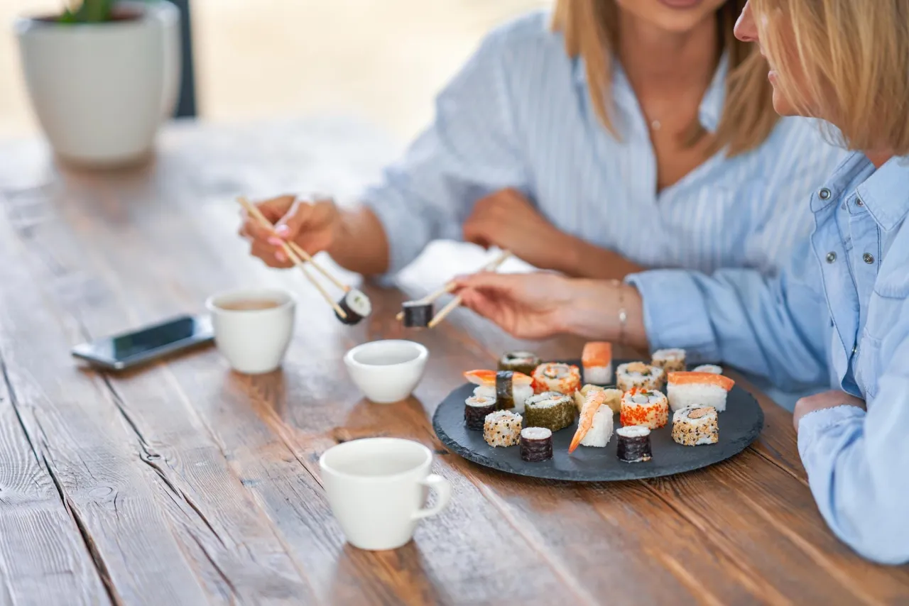 Dwie kobiety jedzą sushi, zastanawiając się, czy można jeść sushi karmiąc piersią. Na stole filiżanki i telefon.