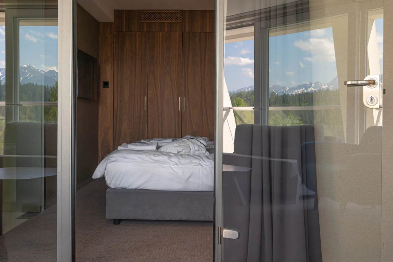 Pok&oacute;j w hotelu Harnaś Wierch, Bukowina Tatrzańska. Ł&oacute;żko z szarym zagł&oacute;wkiem i białym obrusem, drewniana szafa, widok na g&oacute;ry.