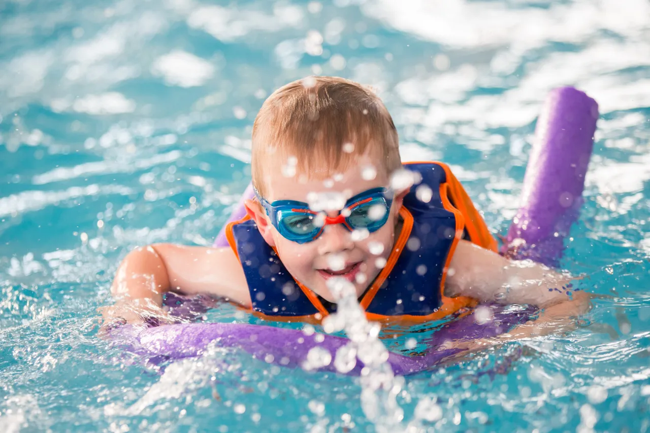 Chłopiec w okularach pływackich i kamizelce ratunkowej uczy się pływać, trzymając się makaronu.