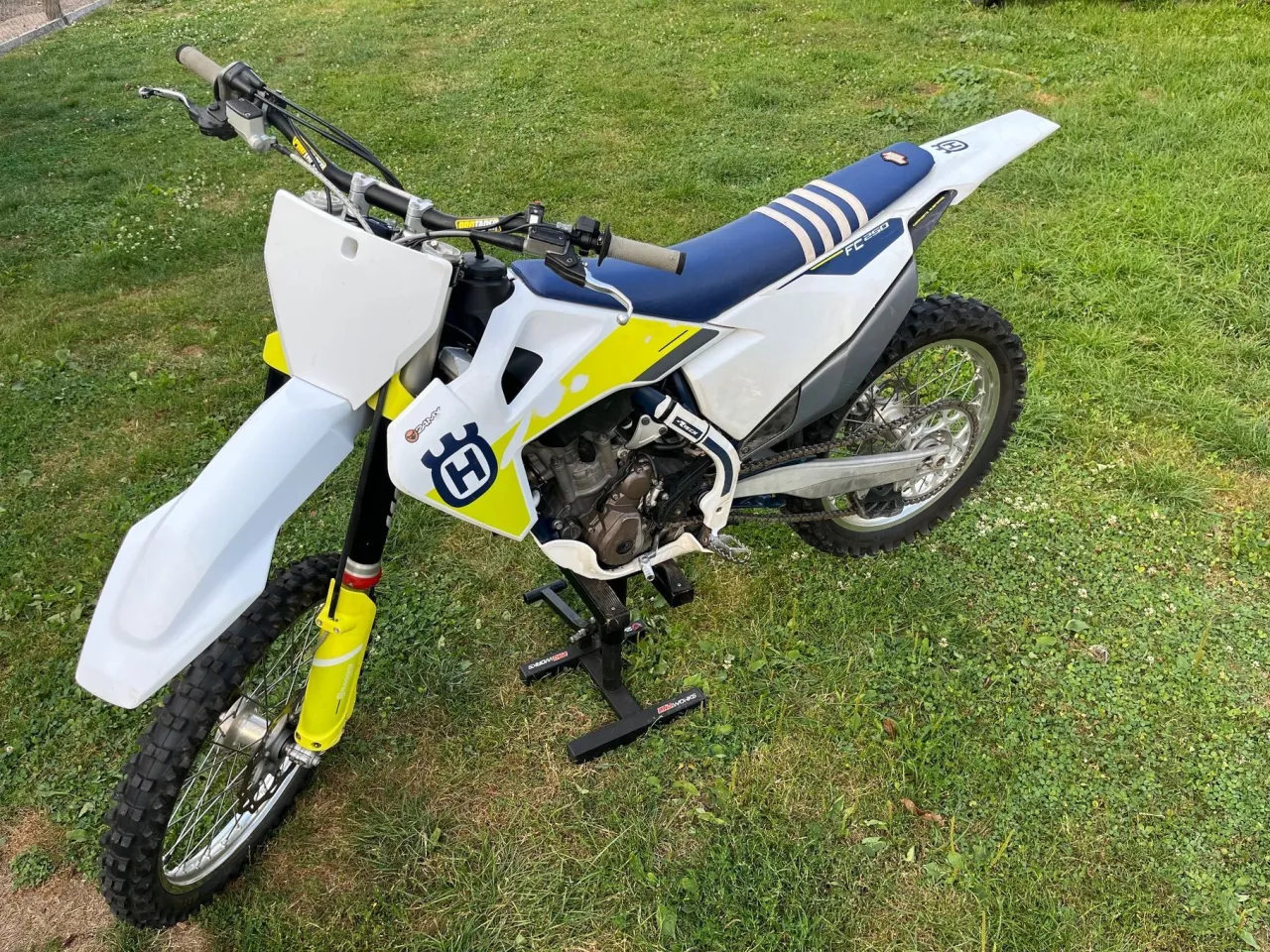 Husqvarna 250 cross na stojaku, gotowy do jazdy po trawiastym terenie. Biało-ż&oacute;łta grafika i niebieskie siedzenie.