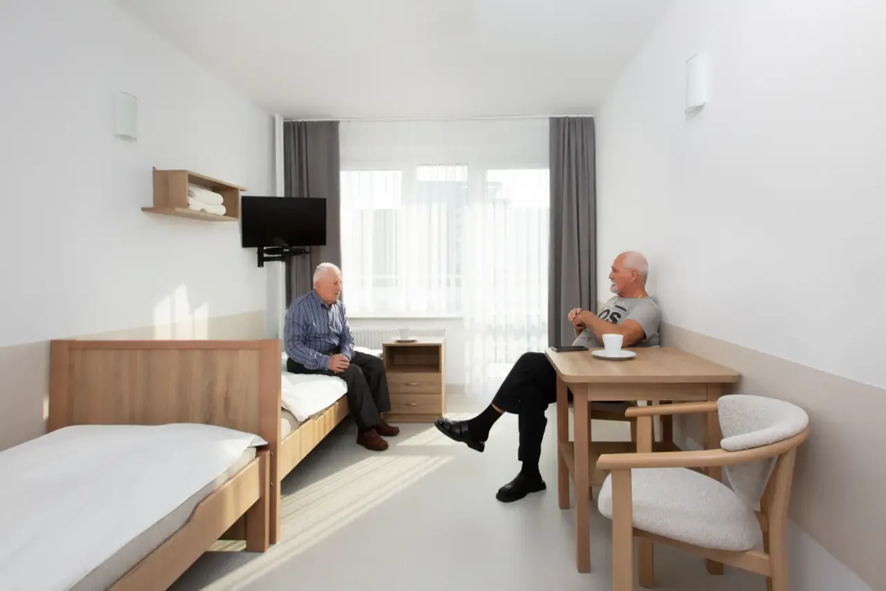 Dwóch starszych panów rozmawia w pokoju sanatorium MSWiA Sopot. Jeden siedzi na łóżku, drugi przy stoliku.