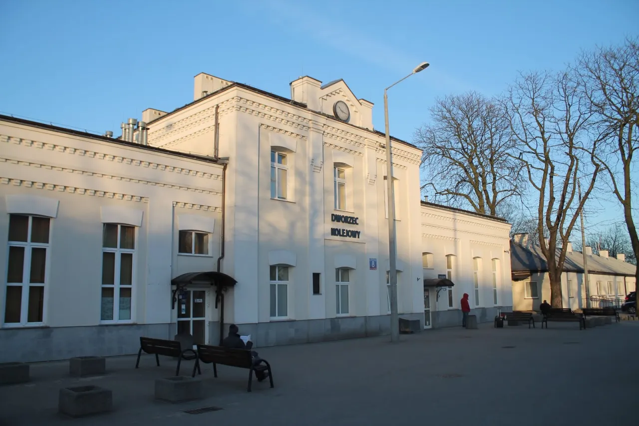 Biały dworzec kolejowy PKP Łódź Widzew z zegarem na fasadzie. Czekający pasażerowie na ławkach.