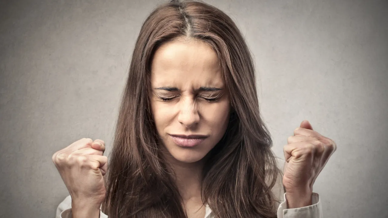 Kobieta z zaciśniętymi pięściami i zamkniętymi oczami, wyrażająca silne emocje. To mogą być objawy choroby, jak ataki złości.