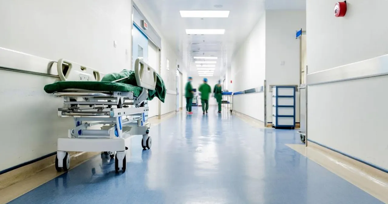 Pusty szpitalny korytarz z wózkiem inwalidzkim, gdzieś ze skręconą kostką. W tle widać niewyraźne sylwetki personelu medycznego.