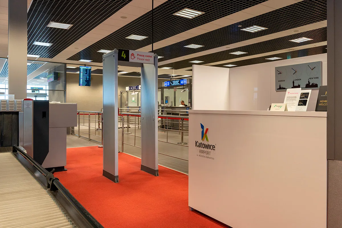 Odprawa na lotnisku w Katowicach: bramki bezpieczeństwa, taśmociąg i recepcja z logo lotniska.