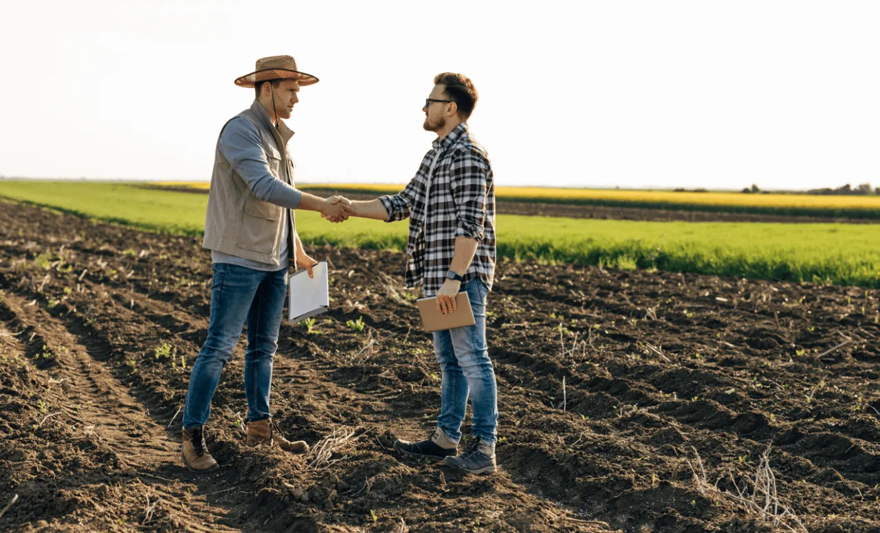 Dwóch mężczyzn w polu, jeden w kapeluszu, ściska sobie dłonie. Może to symbolizować dziedziczenie ziemi rolnej przez nie rolnika.