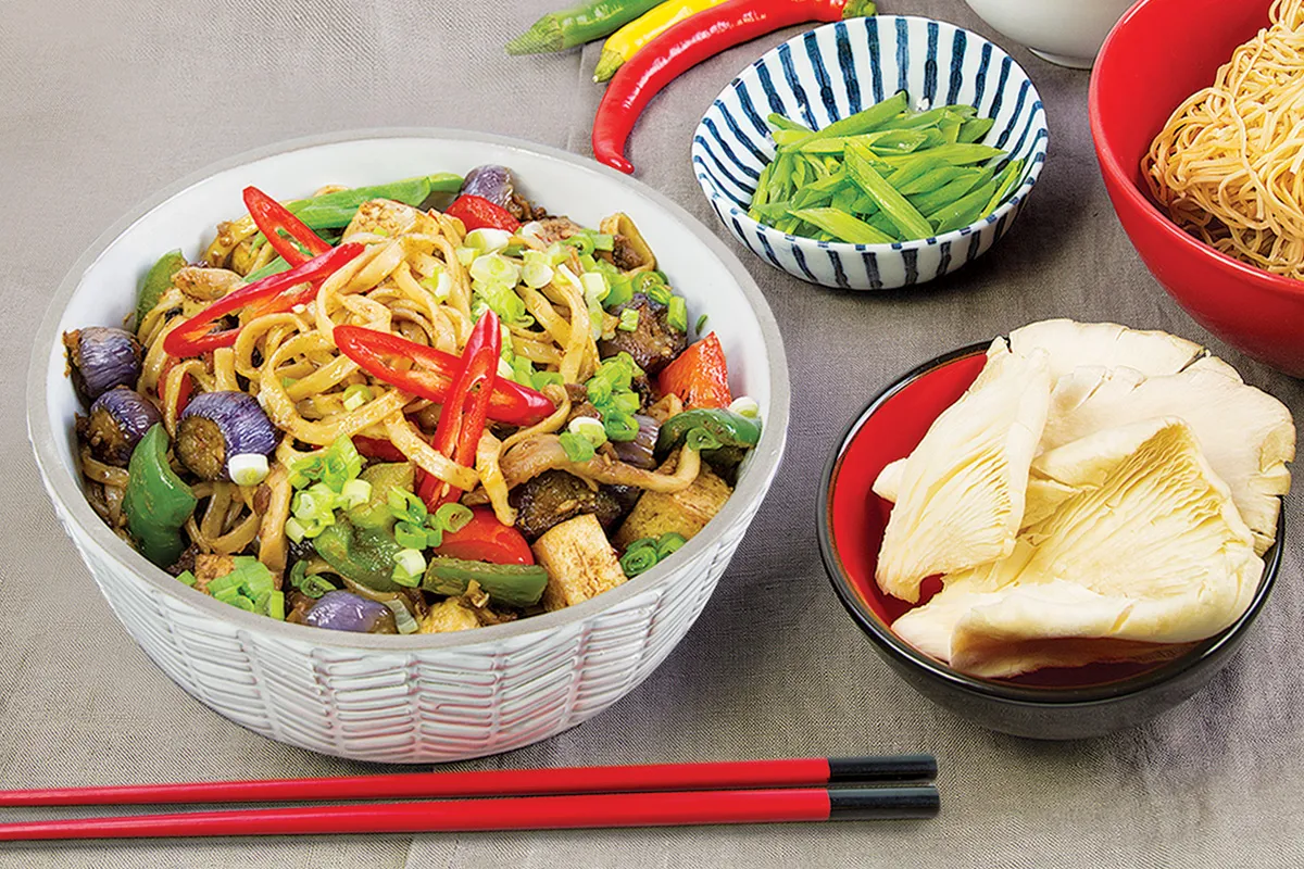 Pyszny makaron chow mein z tofu, papryką, cebulką i ostrymi papryczkami chili. Obok miseczka z zieloną fasolką i grzybami.