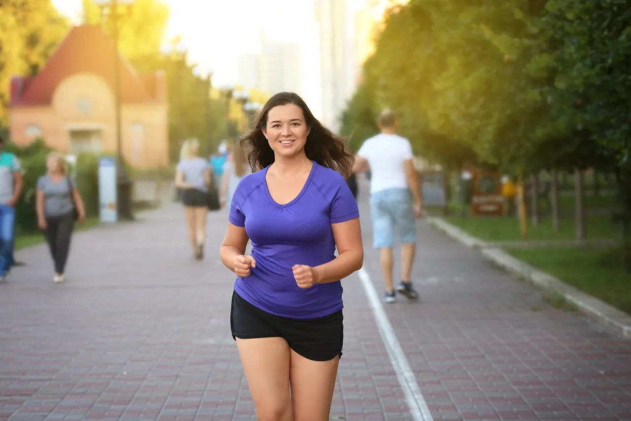 Uśmiechnięta kobieta biegnie po parku, zastanawiając się, ile trzeba biegać żeby schudnąć. W tle inni spacerowicze i drzewa.