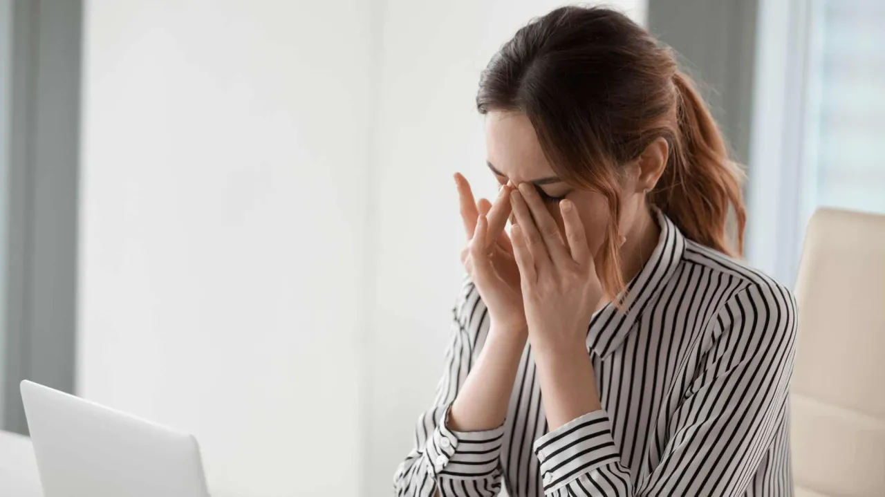 Kobieta zamyka oczy, masując skronie. Widać brak zbieżności objaw&oacute;w, co może wskazywać na zmęczenie lub stres.