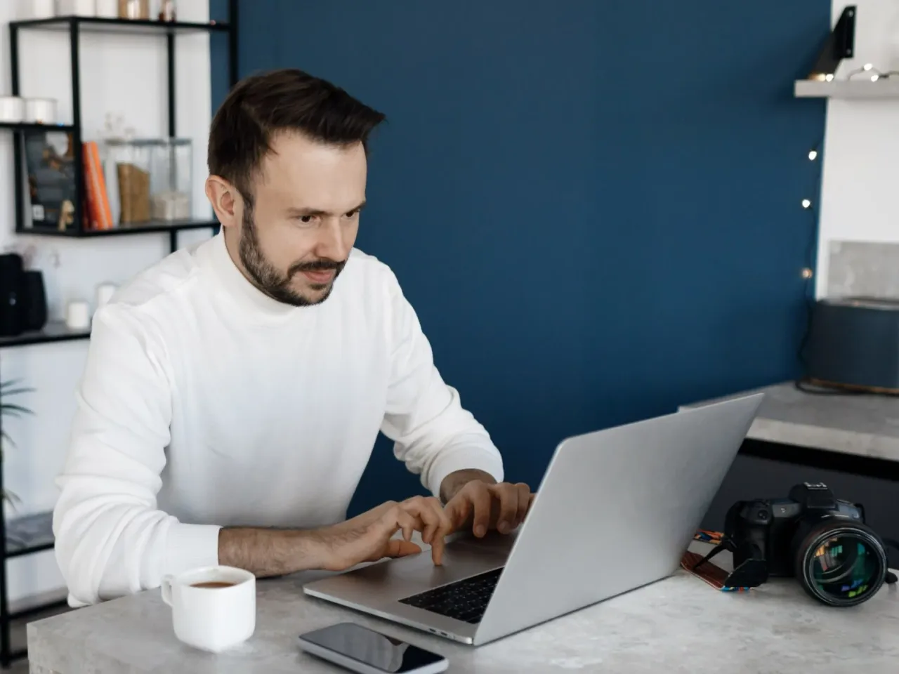 Mężczyzna w białym golfie pracuje na laptopie, obok filiżanka kawy i aparat. Odpowiedź na pytanie: czy podczas rehabilitacji można pracować? Tak!