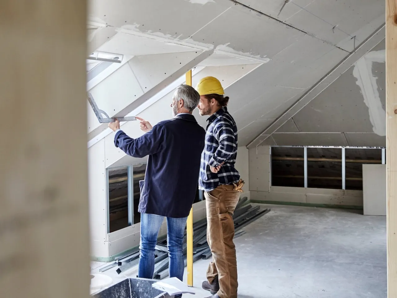 Dwóch mężczyzn planuje adaptację strychu na powierzchnię użytkową. Jeden z nich, w kasku, wskazuje coś na suficie.