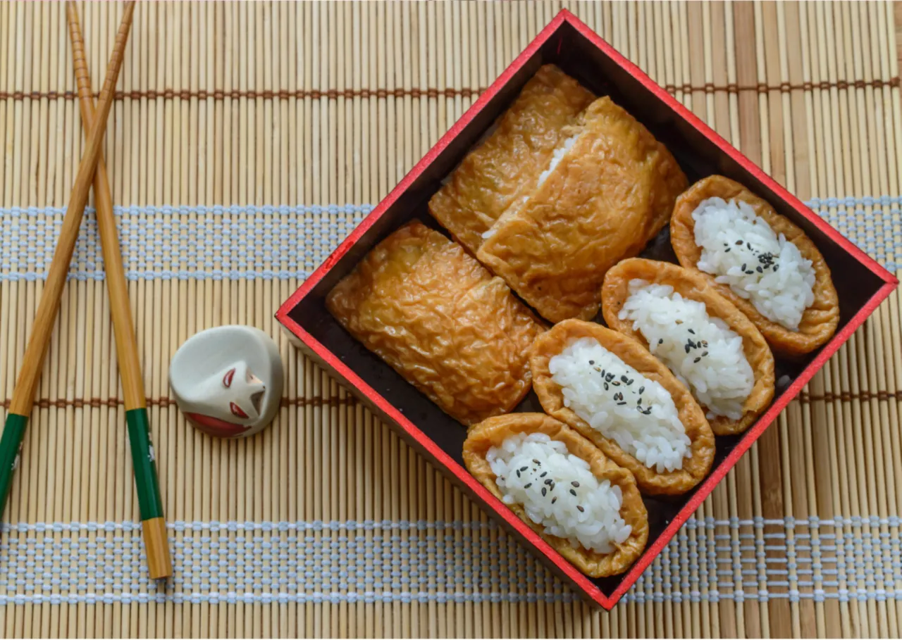 Pyszne inari sushi, czyli ryż w słodkim sosie japońskim, w czerwonym pudełku. Obok pałeczki i ozdobna podstawka.
