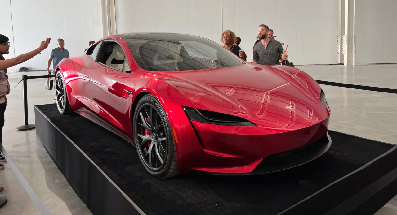 Błyszczący czerwony Tesla Roadster na czarnym cokole. Ludzie oglądają, zastanawiając się, ile kosztuje Tesla Roadster.