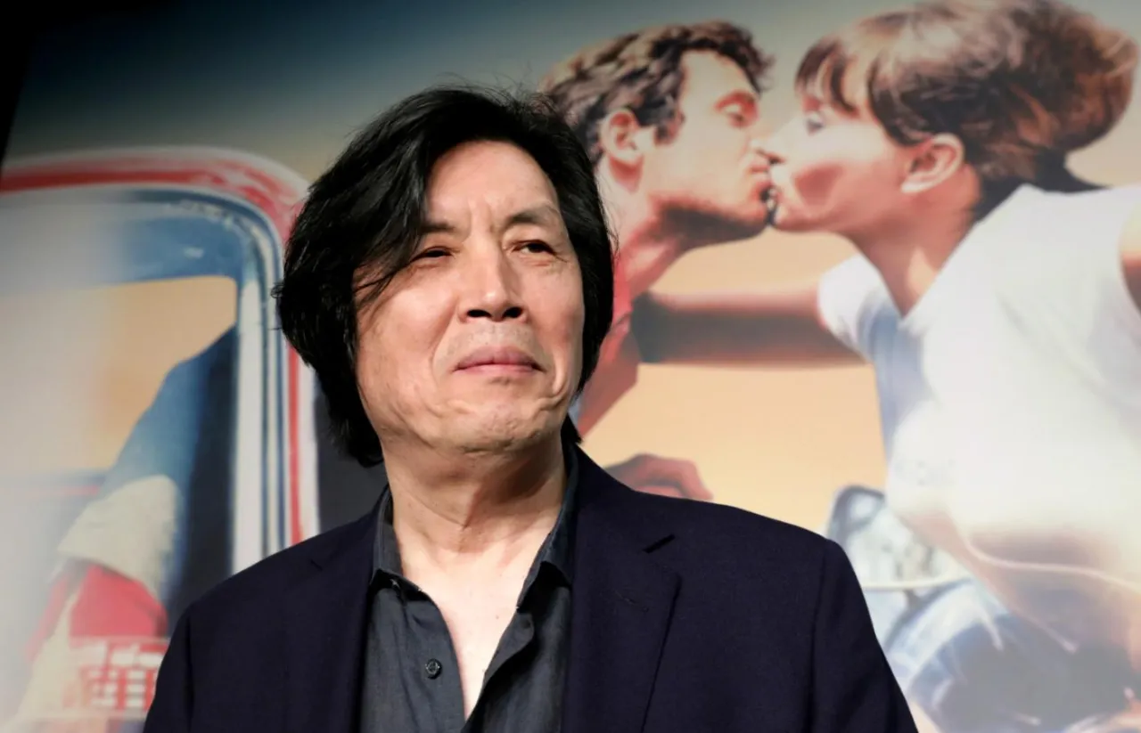 Lee Chang Dong, reżyser, na tle plakatu filmowego z całującą się parą.