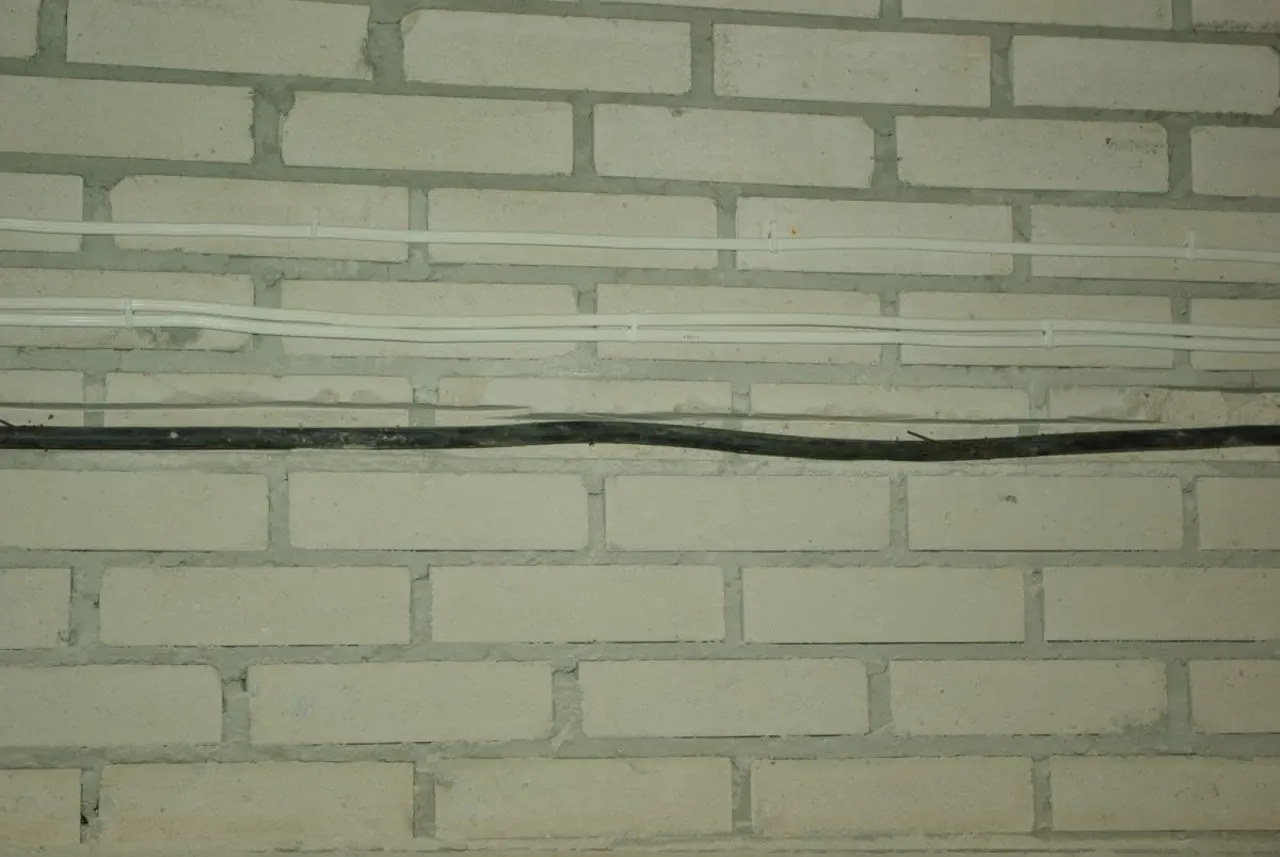 Dwa białe przewody elektryczne i jeden czarny biegną poziomo po jasnej ceglanej ścianie. Ile metr&oacute;w przewodu elektrycznego na dom?