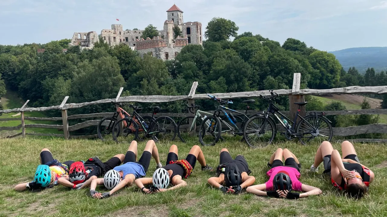 Odpoczynek po rowerowej przygodzie na trasie rowerowej Szlak Orlich Gniazd. Grupa rowerzyst&oacute;w leży na trawie przed ruinami zamku.