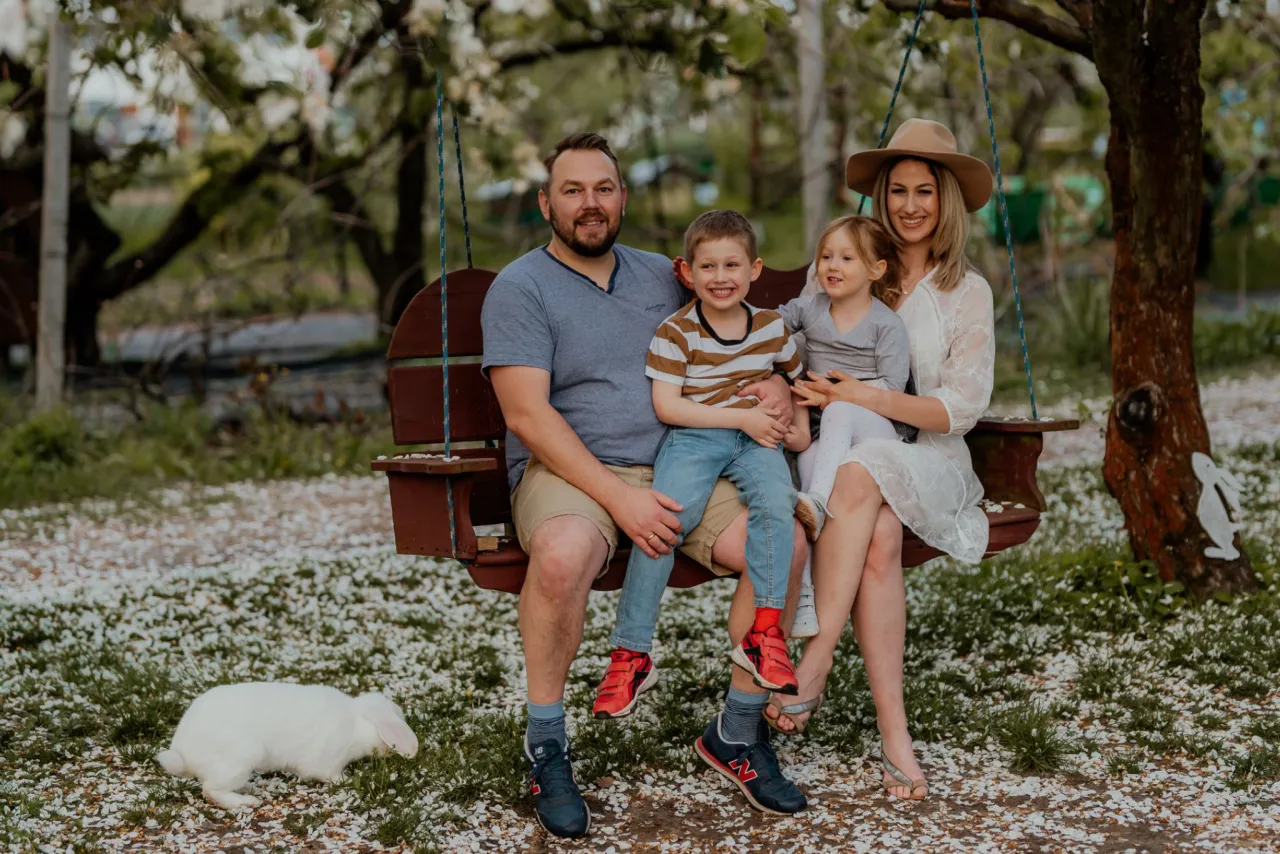Rodzina na huśtawce pod kwitnącym drzewem. Tata z synem na kolanach, mama z c&oacute;rką. W tle biały kr&oacute;lik. Idealne cytaty o tacie.
