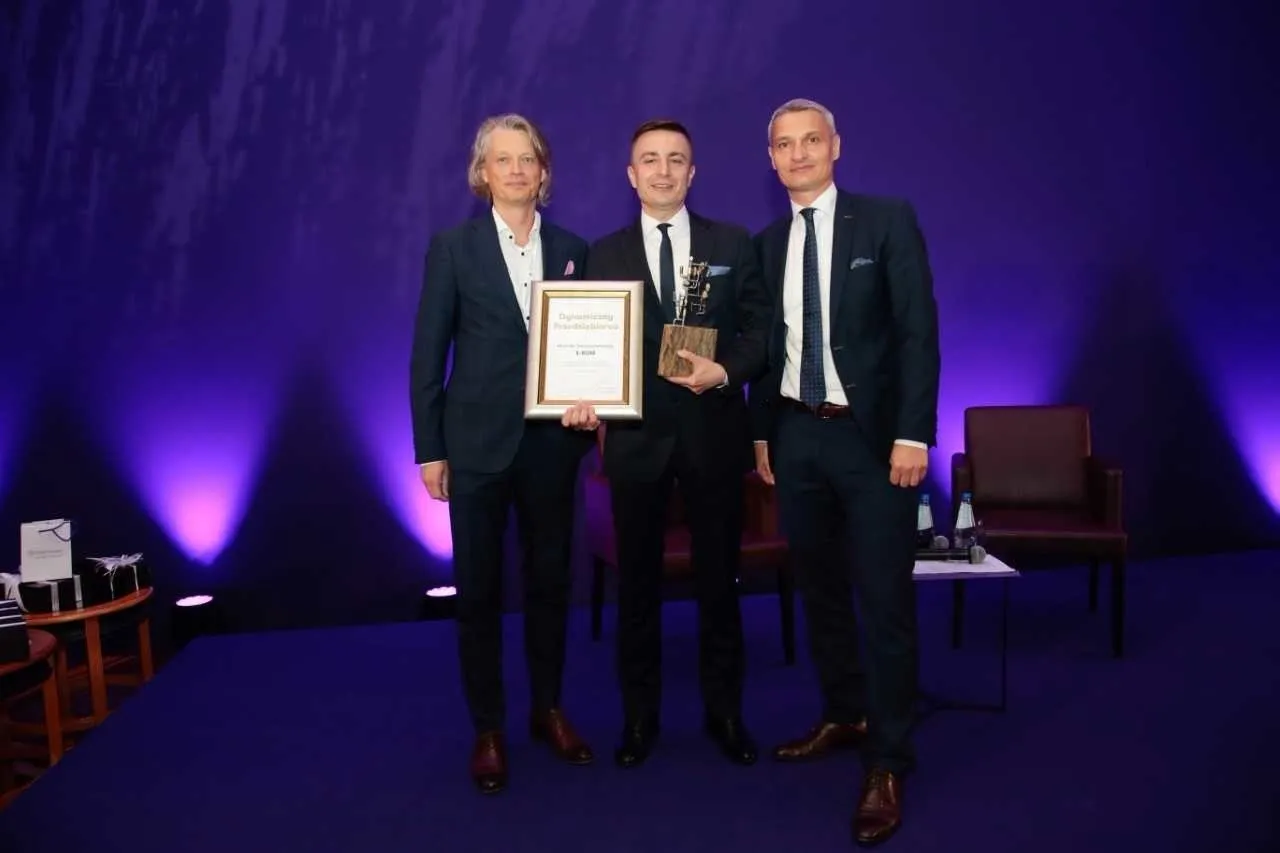 Trzech mężczyzn w garniturach odbiera nagrodę. Na dyplomie widnieje napis 