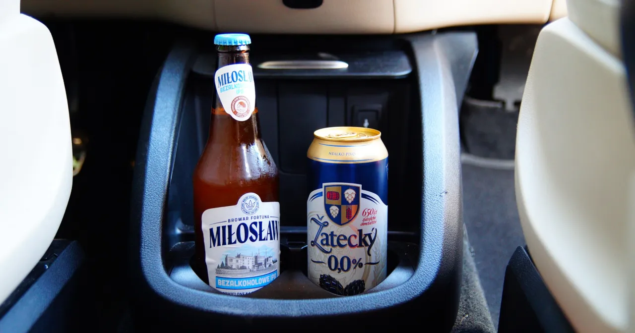 Dwa piwa bezalkoholowe w uchwycie na kubki w samochodzie. Czy można pić piwo bezalkoholowe w miejscu publicznym? Tak, to piwa 0,0%.