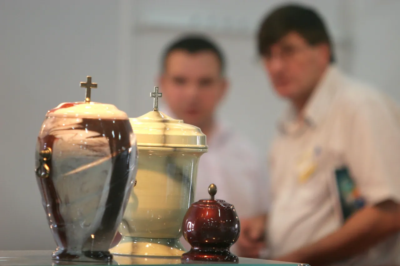 Trzy urny pogrzebowe, dwie z krzyżami, na tle rozmytych postaci. Czy kremacja jest grzechem?