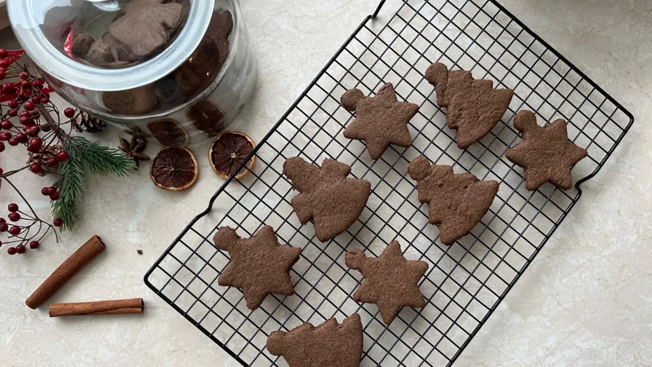 Bezglutenowe pierniki w kształcie gwiazdek i choinek na kratce, obok słoika z piernikami i świątecznych dekoracji.