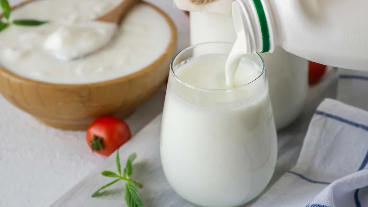 Co to jest ayran? Płynny jogurt nalewany do szklanki, obok miska z jogurtem, pomidorek i listki mięty.