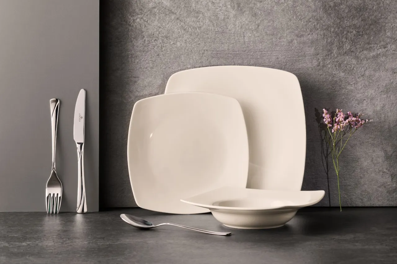 Elegancka ceramika co to? Zestaw kremowych talerzy i miseczki, sztućce i gałązka kwiat&oacute;w na szarym tle.