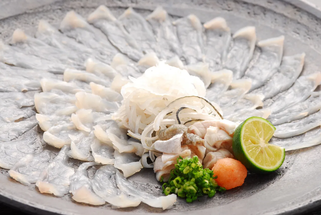 Wykwintne sashimi z ryby fugu, ułożone wachlarzowo, z dodatkiem posiekanej cebulki, pomarańczowej ikry i limonki.