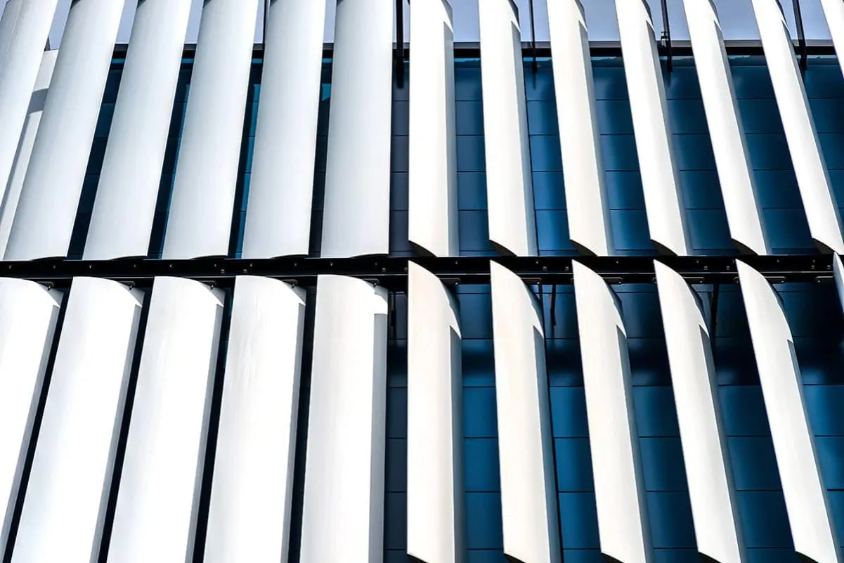 Białe, zakrzywione żaluzje fasadowe zewnętrzne tworzą rytmiczny wzór na ciemnoniebieskiej fasadzie budynku.