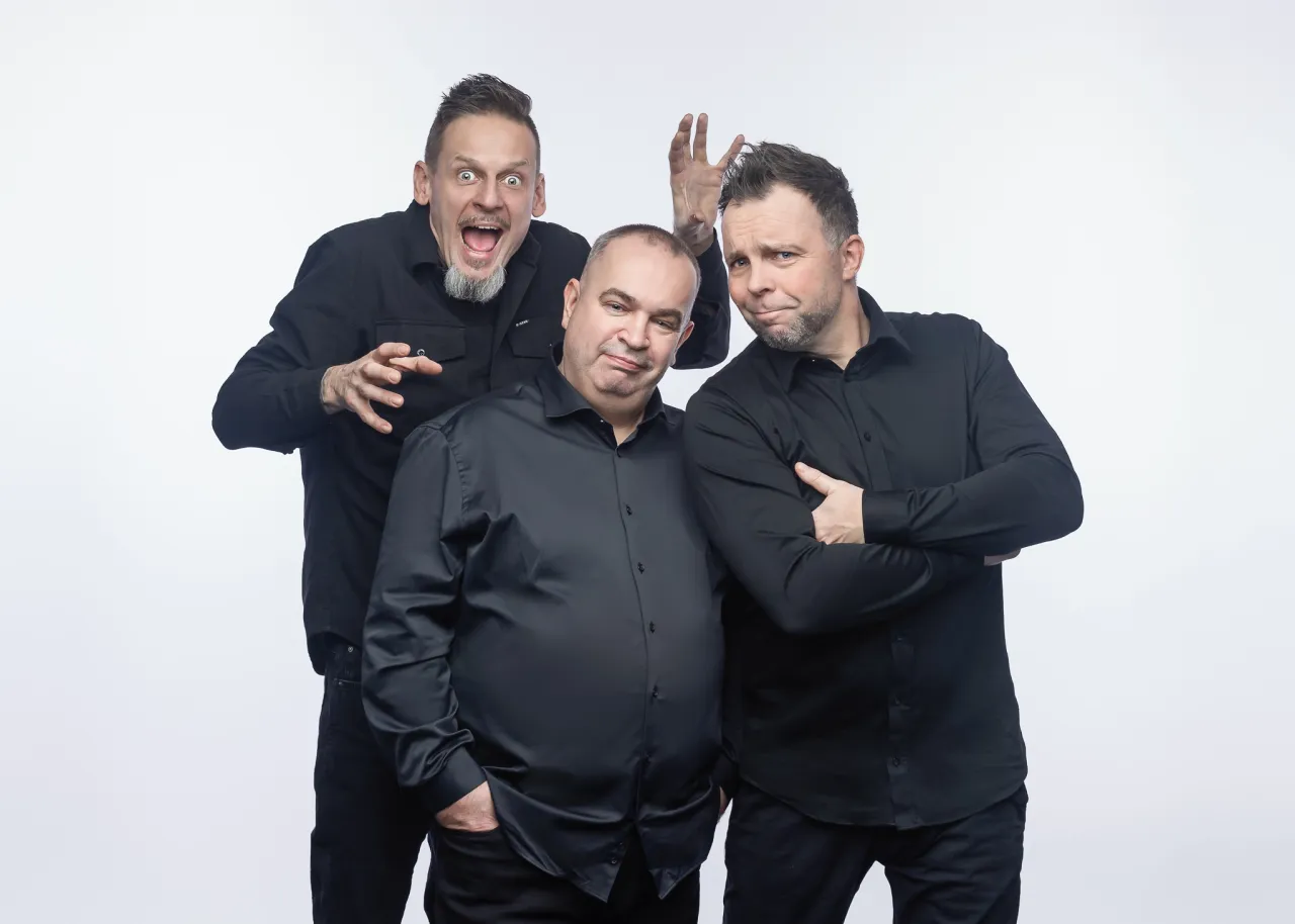 Trzej członkowie kabaretu Ani Mru Mru w czarnych koszulach pozują do zdjęcia. Jeden z nich robi śmieszną minę, drugi ma ręce skrzyżowane na piersi, a trzeci wyciąga ręce.